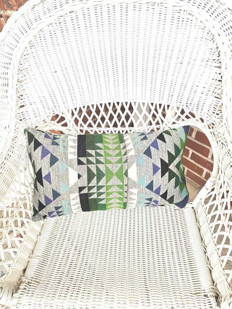 May include: Rectangular decorative pillow with a geometric pattern in shades of gray, green, blue, and white. The pillow is resting on a white wicker chair, creating a contrast of textures and colors. The pillow's design features triangles and other shapes.