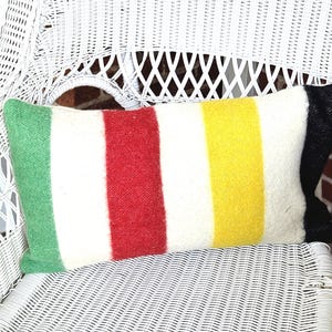 May include: A rectangular pillow with a striped pattern in green, white, red, yellow, and black. The pillow is placed on a white wicker chair, offering a cozy and colorful accent for home decor.