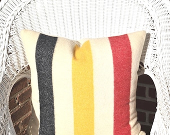 Puede incluir: Un cojín rectangular con fondo crema y rayas verticales negras, amarillas y rojas. El cojín está colocado sobre una silla de mimbre blanca, ofreciendo un acento decorativo y acogedor.