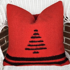 Op de afbeelding: Een rood sierkussen met een zwart boomontwerp en zwarte strepen aan de onderkant. Het kussen is gemaakt van een textuurrijk materiaal en ligt op een witte rieten stoel. Afmetingen circa 40 cm x 40 cm.