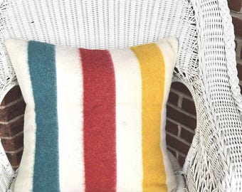 Puede incluir: Un cojín cuadrado con fondo crema y rayas verticales en turquesa, rojo y amarillo. El cojín está sobre una silla de mimbre blanca, con una pared de ladrillo al fondo. No se ven las dimensiones del cojín.