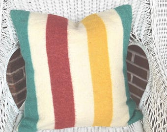 Puede incluir: Un cojín cuadrado con un diseño de rayas en colores turquesa, rojo, amarillo y crema. El cojín está hecho de una tela suave y texturizada y está colocado sobre una silla de mimbre blanca.