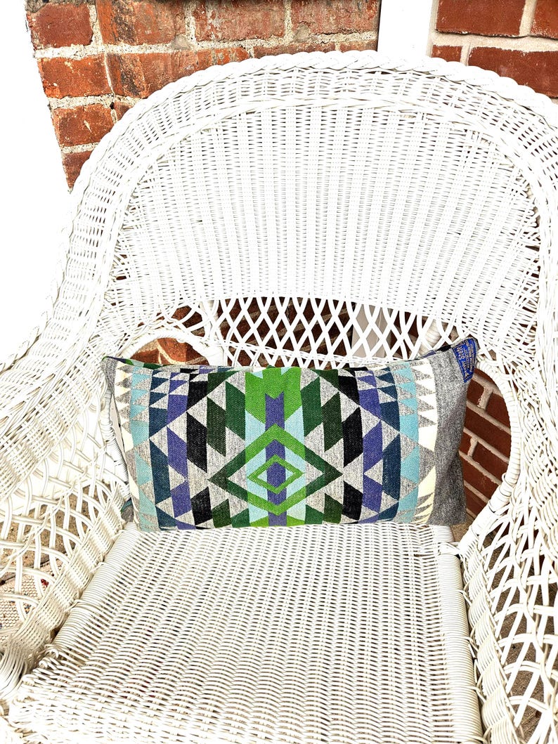 May include: A rectangular decorative pillow with a geometric pattern in shades of blue, green, and gray. The pillow is placed on a white wicker chair, with a brick wall in the background.