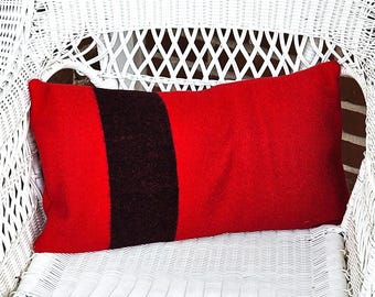 Puede incluir: Un cojín rectangular con una base de tela roja vibrante y una única franja vertical negra ancha situada hacia el lado izquierdo. El cojín tiene una textura suave similar al fieltro y se muestra horizontalmente sobre una silla de mimbre blanca.