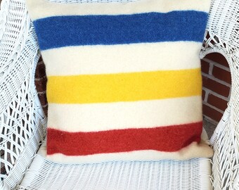 Puede incluir: Un cojín cuadrado con rayas horizontales azules, amarillas y rojas sobre un fondo crema. El cojín está hecho de una tela gruesa y texturizada y está colocado sobre una silla de mimbre blanca.