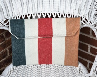 May include: A rectangular felt clutch with a striped pattern in teal, white, red, and tan. The clutch is folded over at the top and secured with a white string. The clutch is resting on a white wicker chair.