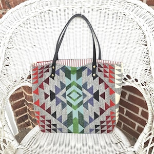 May include: A gray tote bag with a geometric pattern in red, blue, green, and black. The bag has black leather handles and is sitting on a white wicker chair. The interior lining has a red and white striped pattern.