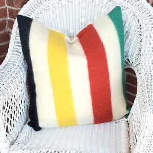 May include: A square pillow with a cream-colored background and vertical stripes in black, yellow, red, and green. The pillow is made of a soft, textured material and is placed on a white wicker chair.