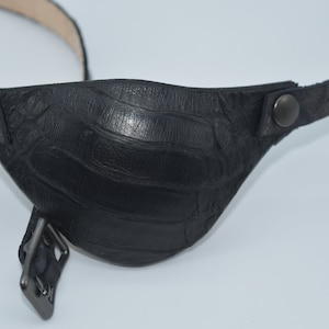 May include: Black leather eye patch with a crocodile pattern and a metal buckle. The patch is attached to a black leather strap with a metal button.