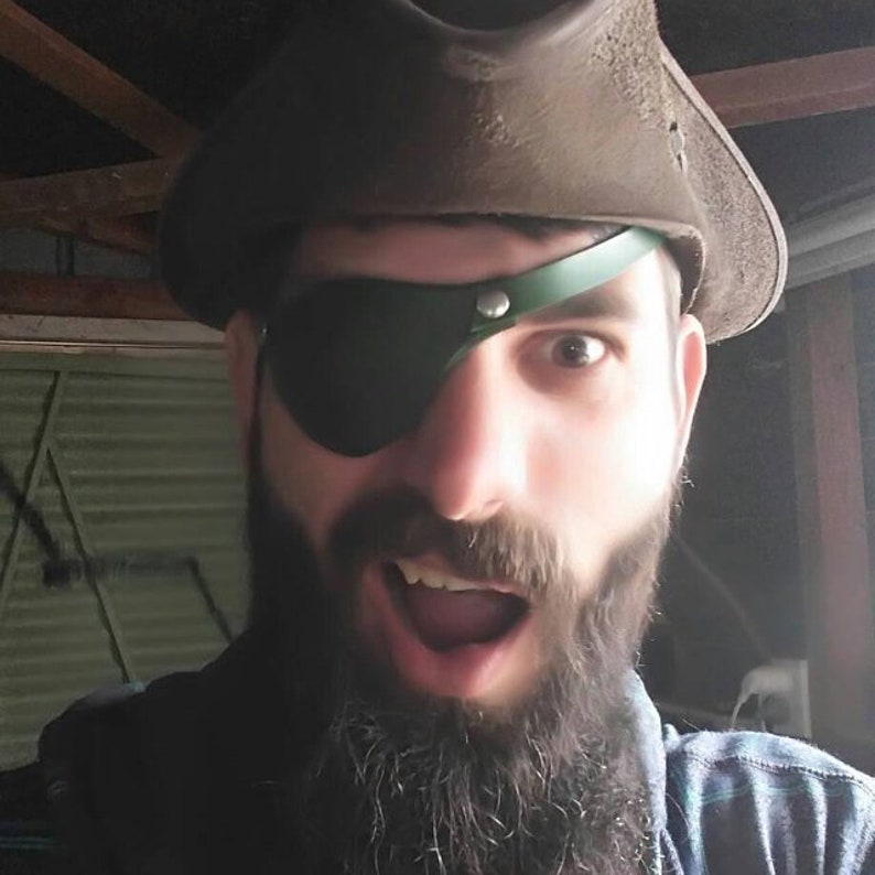 May include: A man with a beard and an eye patch is wearing a brown leather pirate hat. He is looking at the camera with a surprised expression.
