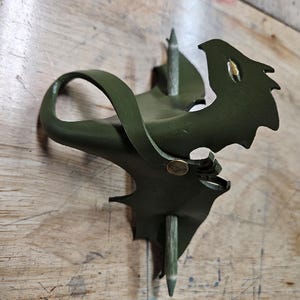Op de afbeelding: Handgemaakte haarspeld van olijfgroen leer in de vorm van een draak. De draak is gedetailleerd met een messing klinknagel en een houten stok om het haar vast te zetten. De vleugels en staart van de draak zijn zichtbaar, op een houten ondergrond.
