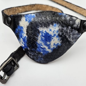 May include: A black leather eye patch with a blue and white tie-dye pattern. The patch is attached to a black leather strap with a metal buckle.