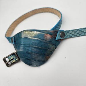 May include: A blue leather eye patch with a gold and brown pattern. The patch has a strap with a buckle and a metal stud.