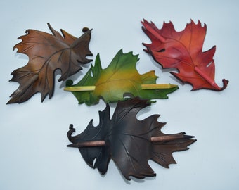May include: Four leather leaf hair clips in different colours: brown, green, yellow, and red. Each clip has a wooden or copper stick that goes through the leaf.