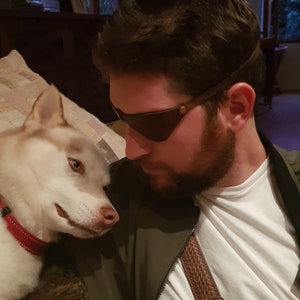 May include: A man with a beard wearing a brown leather eye patch and a white t-shirt is looking at a white and brown husky dog.