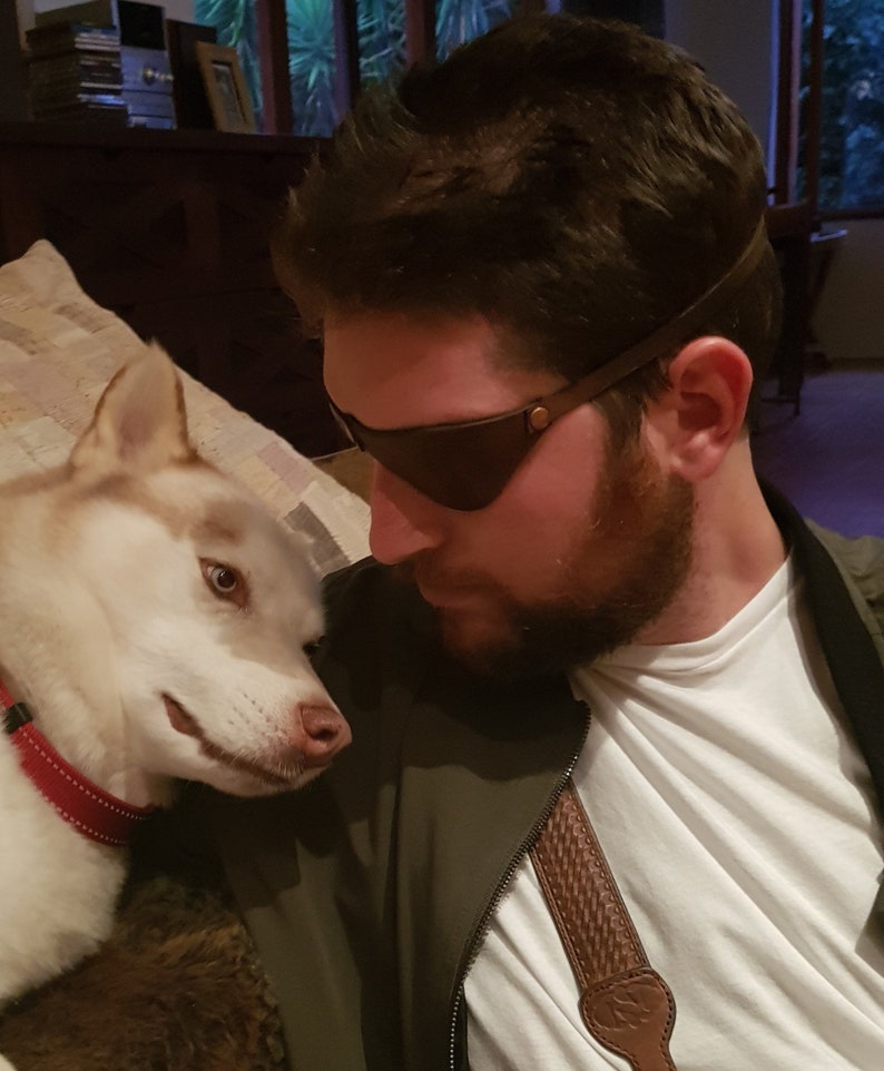 May include: A man with a beard wearing a brown leather eye patch and a white t-shirt is looking at a white and brown husky dog.
