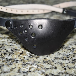 Peut inclure: Un cache-œil en cuir noir avec une sangle et plusieurs trous. Le cache-œil est posé sur un plan de travail en granit.