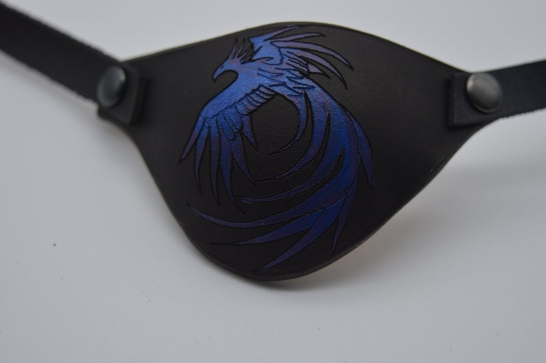 Puede incluir: Parche para el ojo de cuero negro con un dise&ntilde;o de f&eacute;nix azul y morado.