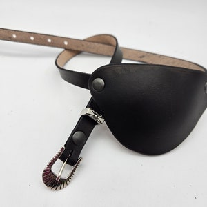 May include: A black leather belt with a silver buckle and a decorative flap. The belt is designed for a cowboy or western style.