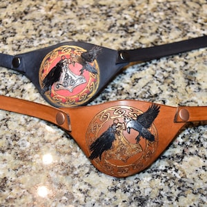 May include: Two leather eye patches with a design of two ravens holding a hammer in a circle. One eye patch is black leather with a red background and gold accents. The other eye patch is brown leather with a gold background and gold accents.