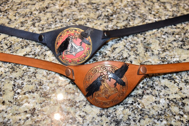 May include: Two leather eye patches with a design of two ravens holding a hammer in a circle. One eye patch is black leather with a red background and gold accents. The other eye patch is brown leather with a gold background and gold accents.