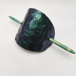 May include: A green and black hair clip with a textured, scaly design. The clip is made of leather and has a wooden stick.