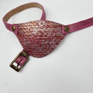 May include: A pink leather eye patch with a gold and white patterned design. The patch is attached to a pink leather strap with a brass buckle.