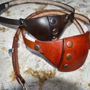 May include: Two leather eye patches with adjustable straps. One is brown with silver accents and the other is orange with copper accents.