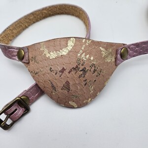 May include: A pink leather eye patch with a gold patterned design. The eye patch is attached to a pink leather strap with a buckle.