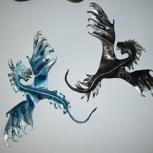 May include: Two metal dragon wall sculptures. One is blue and silver, the other is black and silver. Both have detailed wings and tails, and are mounted on a white wall. Fantasy decor.