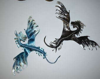 Puede incluir: Dos esculturas de pared de dragón de metal. Una es azul y plateada, la otra negra y plateada. Ambas tienen alas y colas detalladas y están montadas en una pared blanca. Decoración de fantasía.