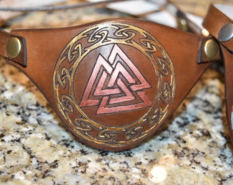 Op de afbeelding: Een bruin leren ooglapje met een goud- en koperkleurig Valknut-symbool in het midden. Het symbool wordt omringd door een gouden Keltisch knoopontwerp.