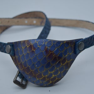 Peut inclure: Un cache-œil en cuir bleu avec un motif de peau de serpent et une sangle en cuir bleu avec une boucle.