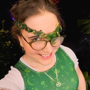 Peut inclure: Une personne porte un haut vert et blanc avec un motif sur le thème de la nature et un serre-tête décoratif avec des feuilles vertes et des accents bruns. Elle porte également des lunettes avec une monture marron clair.