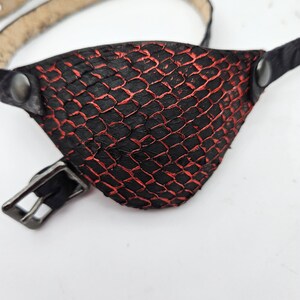 May include: A black and red eye patch with a reptile scale pattern. The eye patch is attached to a thin, light brown leather strap with a buckle and metal rivets. A unique accessory.