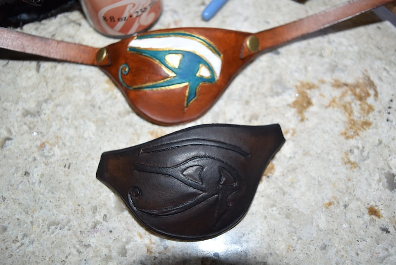 Op de afbeelding: Twee leren ooglapjes met een Egyptisch oog van Horus-ontwerp. E&eacute;n is bruin met een blauw en gouden oog, de andere is zwart met een zwart oog.