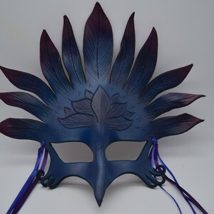 May include: A blue and purple leather masquerade mask with a lotus flower design and feather-like accents. The mask has two purple ribbons attached to the sides.