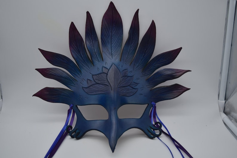 May include: A blue and purple leather masquerade mask with a lotus flower design and feather-like accents. The mask has two purple ribbons attached to the sides.