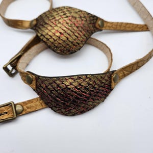 May include: A pair of brown leather eye patches with gold accents and a textured surface. The eye patches are attached to a brown leather strap.