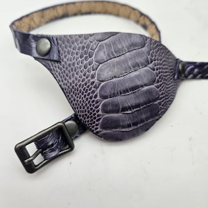May include: A purple leather eye patch with a textured, reptile-like pattern. The eye patch has a strap and buckle for adjustment, and a strap that goes around the head. The eye patch is designed to cover one eye.