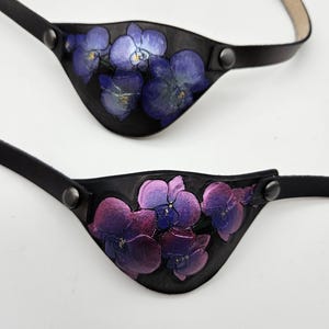 May include: Two black leather eye patches, each with a floral design. One displays blue orchids, the other purple. Both have black straps and snap closures for a secure fit.