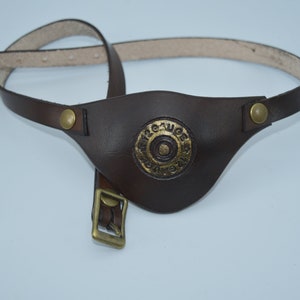 K&ouml;nnte beinhalten: Ein braunes Leder-Augenpflaster mit einer goldenen Metallschnalle und einem kreisf&ouml;rmigen Design mit dem Text "12 Gauge" in der Mitte.