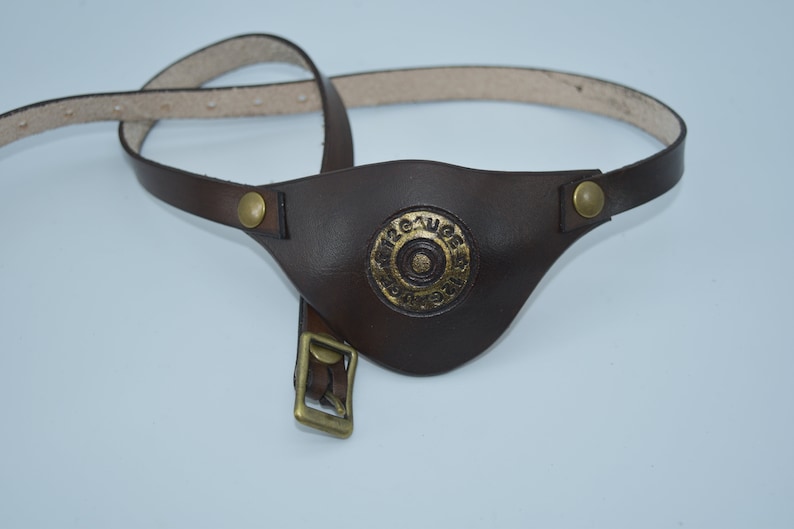 K&ouml;nnte beinhalten: Ein braunes Leder-Augenpflaster mit einer goldenen Metallschnalle und einem kreisf&ouml;rmigen Design mit dem Text "12 Gauge" in der Mitte.