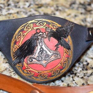 May include: A black leather eye patch with a red and gold design. The design features a hammer, two ravens, and a Celtic knot.
