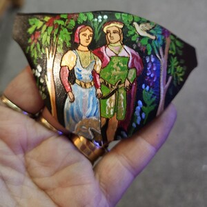 Op de afbeelding: Een handgeschilderd leren ornament dat een stel in een bosrijke omgeving voorstelt. De vrouw draagt een blauwe jurk en de man draagt een groene en rode tuniek. De achtergrond is een donkergroen bos met bomen en bloemen.