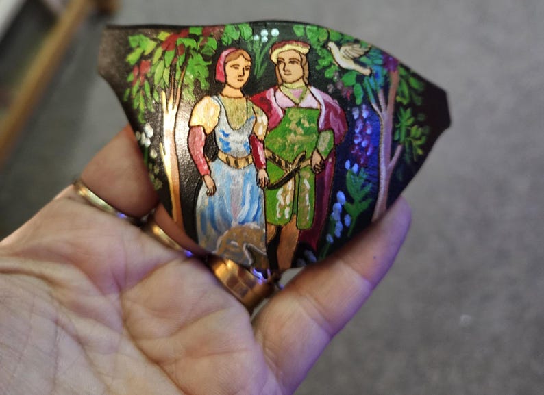 Op de afbeelding: Een handgeschilderd leren ornament dat een stel in een bosrijke omgeving voorstelt. De vrouw draagt een blauwe jurk en de man draagt een groene en rode tuniek. De achtergrond is een donkergroen bos met bomen en bloemen.