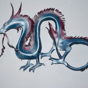 May include: A blue and purple metal dragon sculpture with intricate details. The dragon has a long, serpentine body, sharp claws, and a detailed head with a long tongue. The sculpture is mounted on a white surface.
