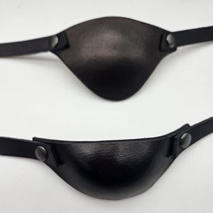 May include: Two black leather eye patches with adjustable straps. Each patch is curved to fit the eye and secured with a dark metal snap. The leather has a slightly textured appearance.