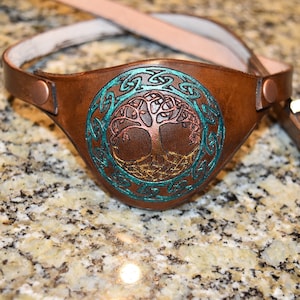 May include: A brown leather eye patch with a Celtic knot design and a tree of life motif in teal, gold, and copper colors.