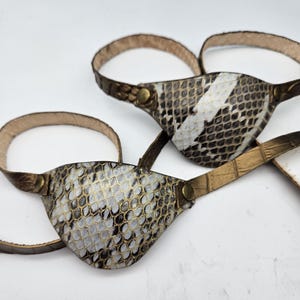 Peut inclure: Deux cache-œils présentant un motif texturé en peau de serpent dans des tons de blanc, gris et noir. Chaque cache-œil est fixé à une fine sangle réglable en matériau bronze métallique, sécurisée par de petits rivets métalliques couleur laiton.